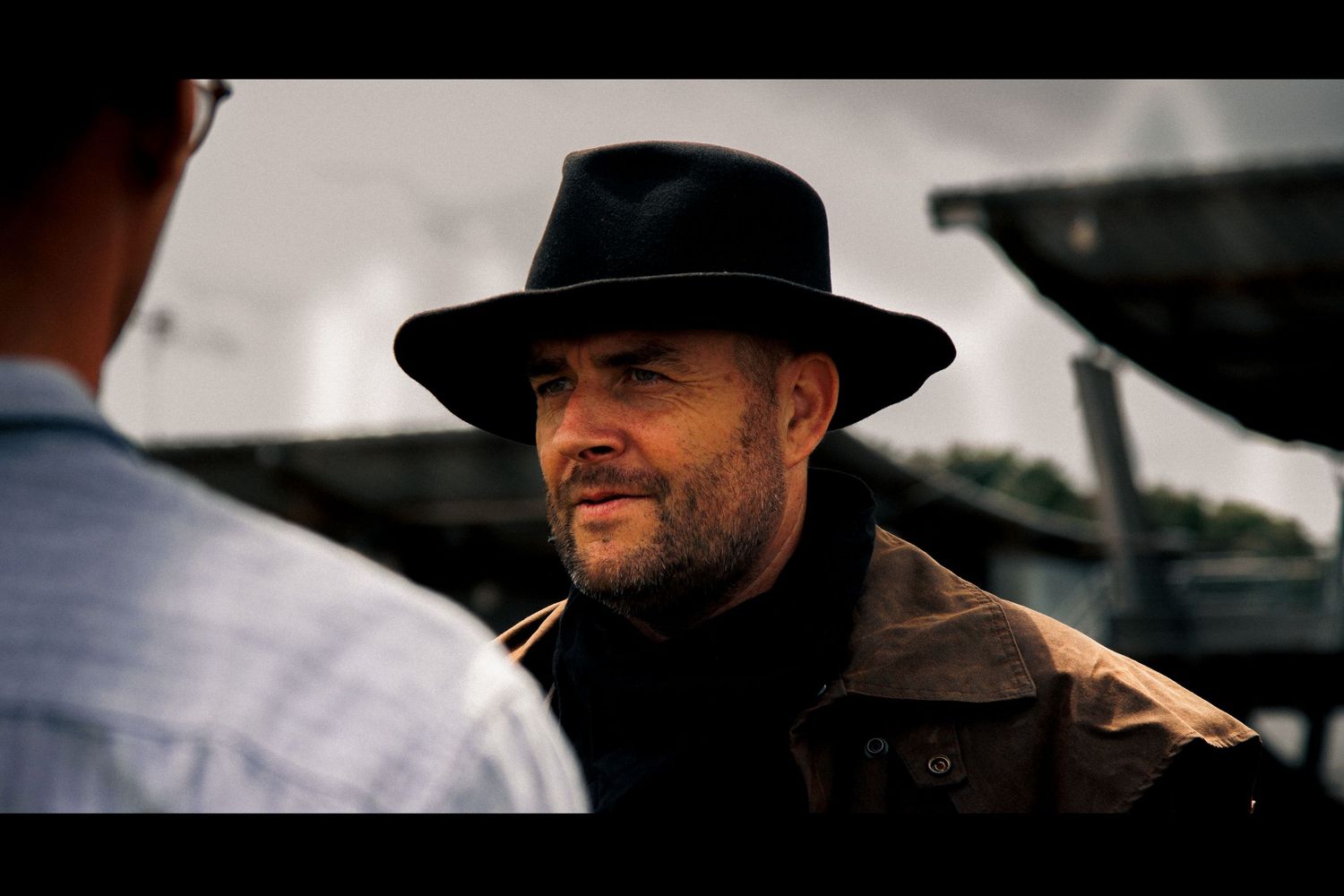 Person in black hat and coat speaks during dramatic western scene.