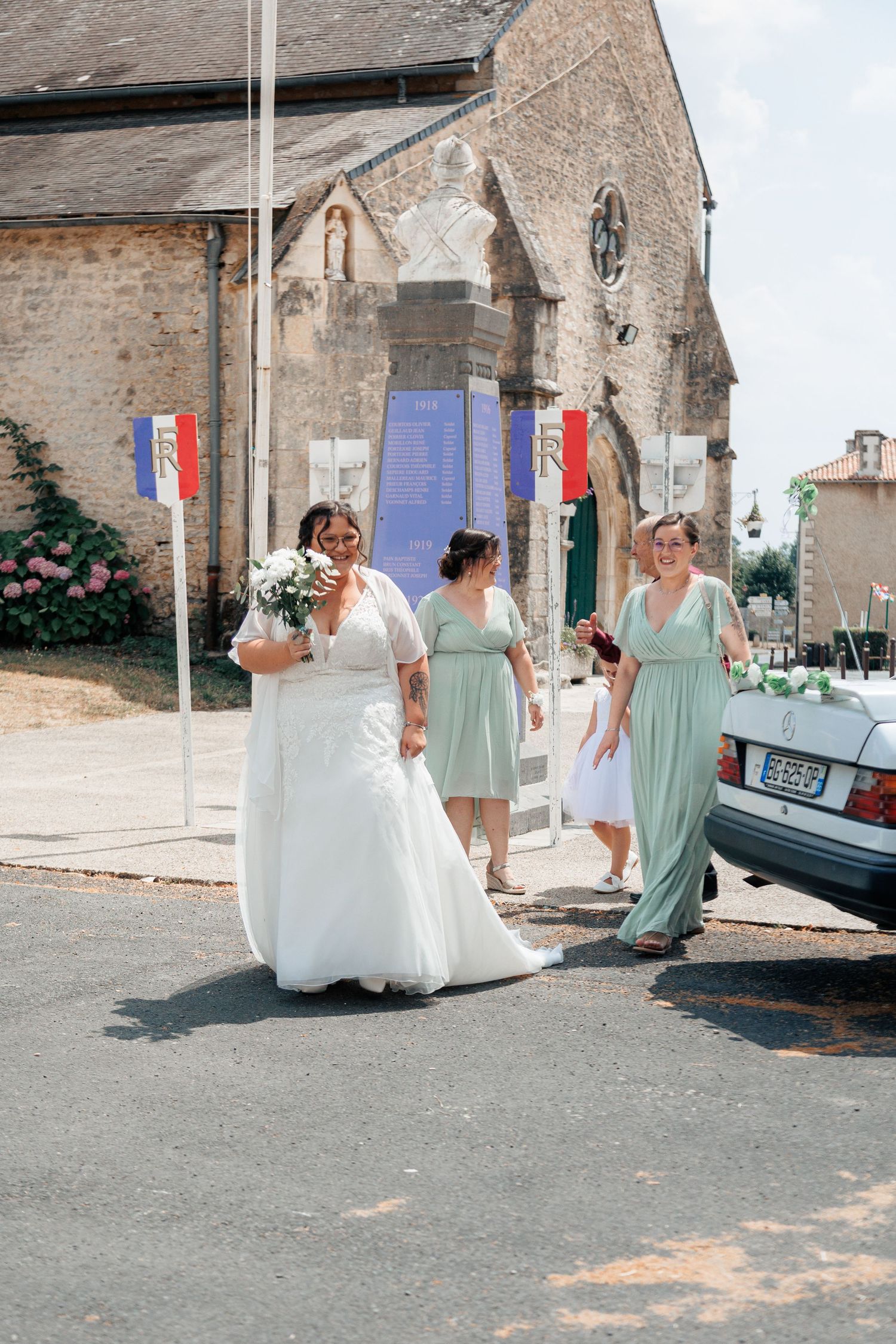 Bride in white gown walks with bridesmaids in mint dresses outside a French church on a sunny wedding day.