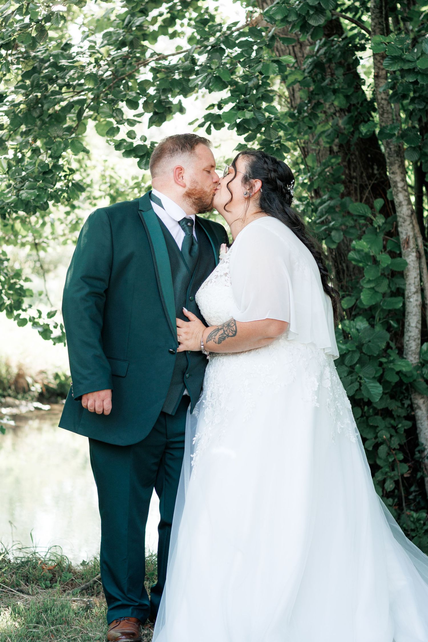 Newlyweds share romantic kiss under green trees near water in wedding portraits.
