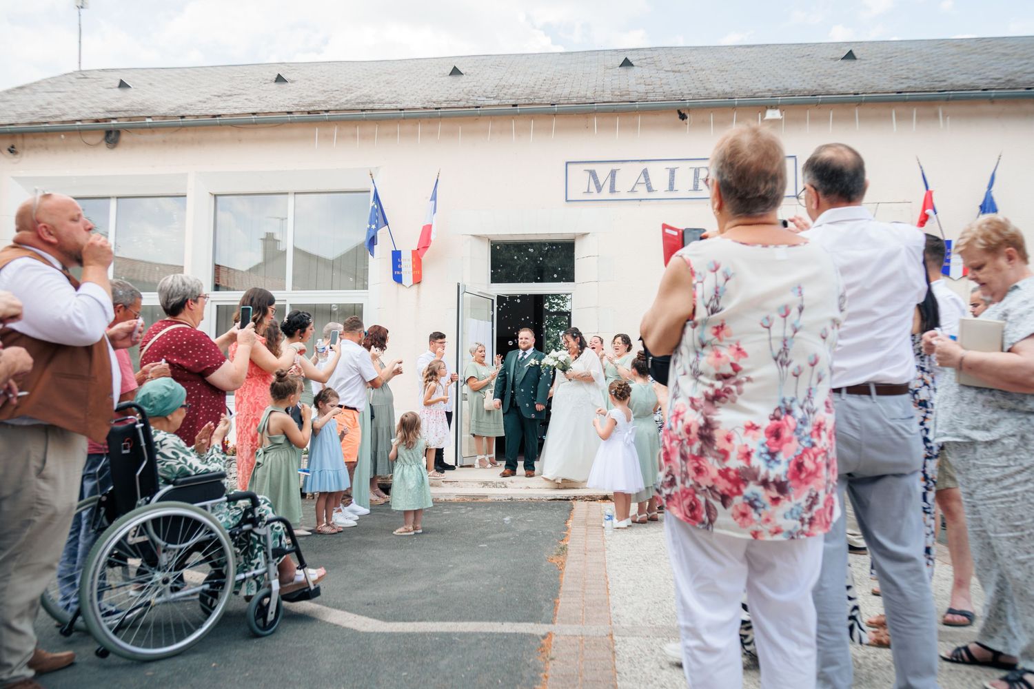 Newlyweds exiting French mairie greeted by cheering guests throwing confetti on sunny wedding day.
