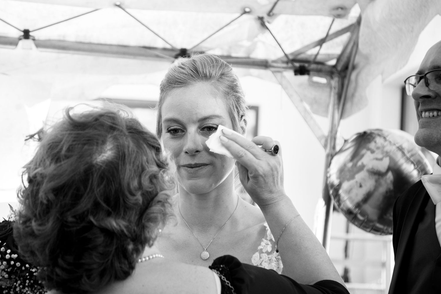 Black and white photo of bride having makeup touched up by two women during wedding day preparations.