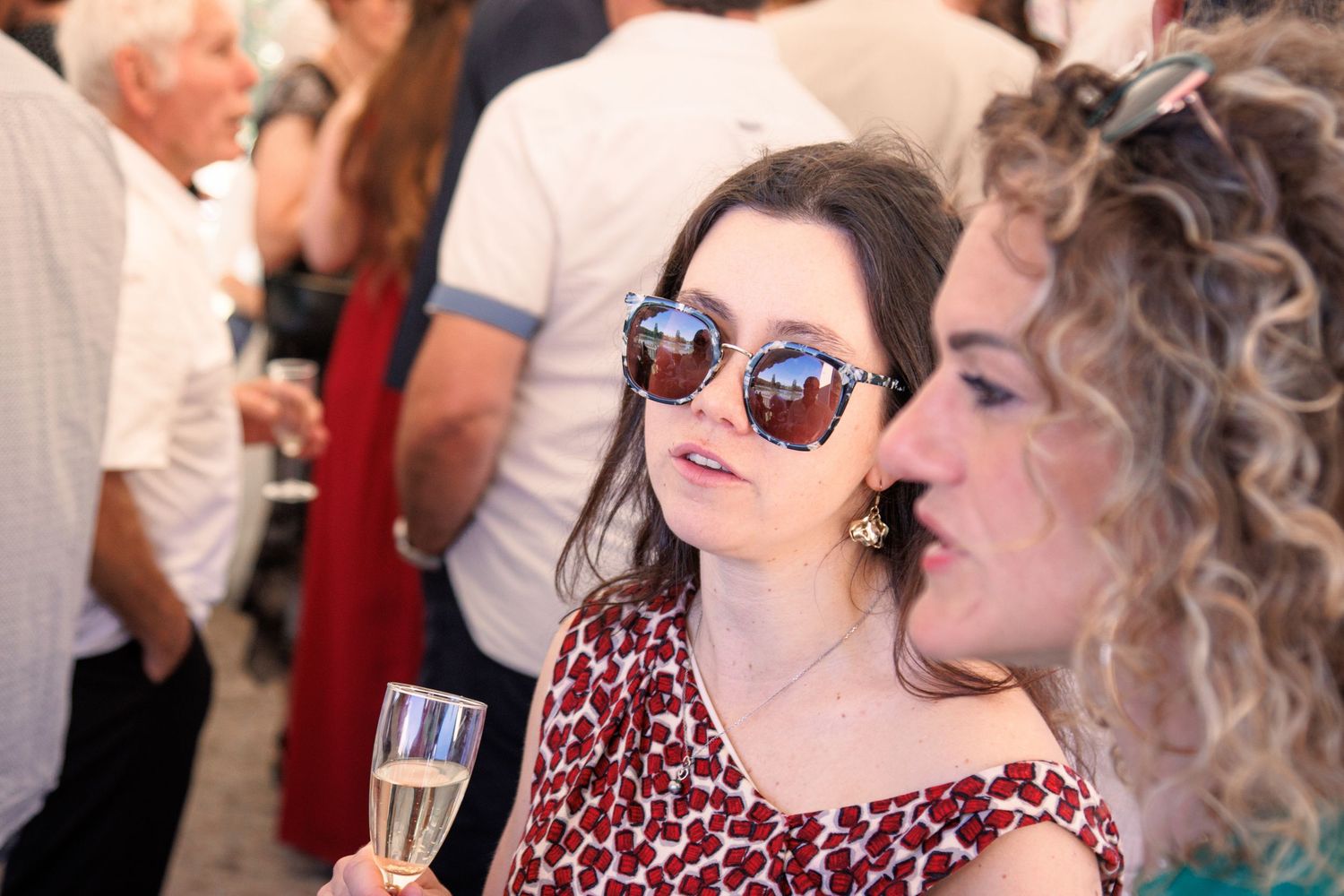 Two young women at a wedding reception, one wearing sunglasses holding a champagne glass.
