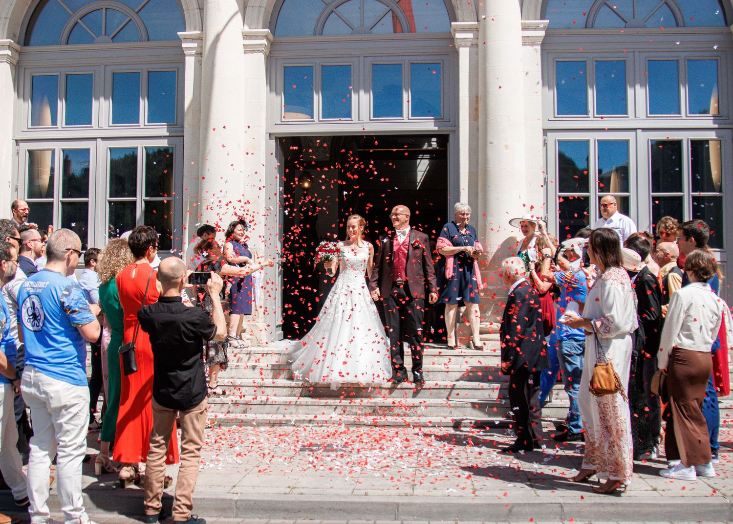 Newlyweds exit a grand white building as guests shower them with colorful confetti at their wedding.