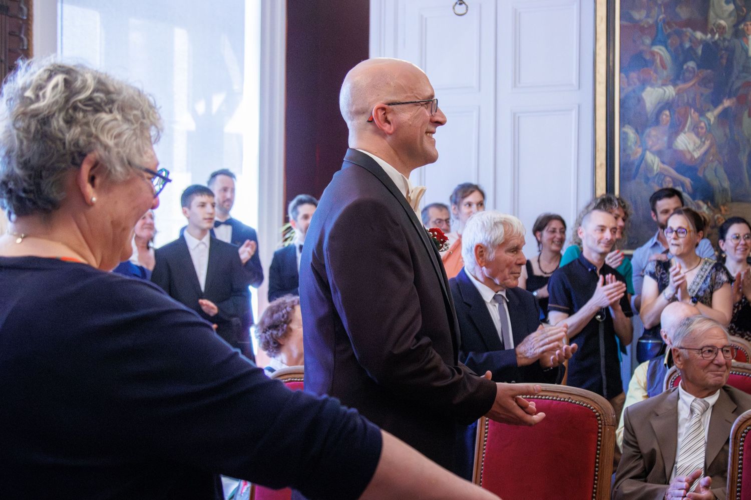Bald man in dark suit smiles at guests during elegant indoor wedding reception.
