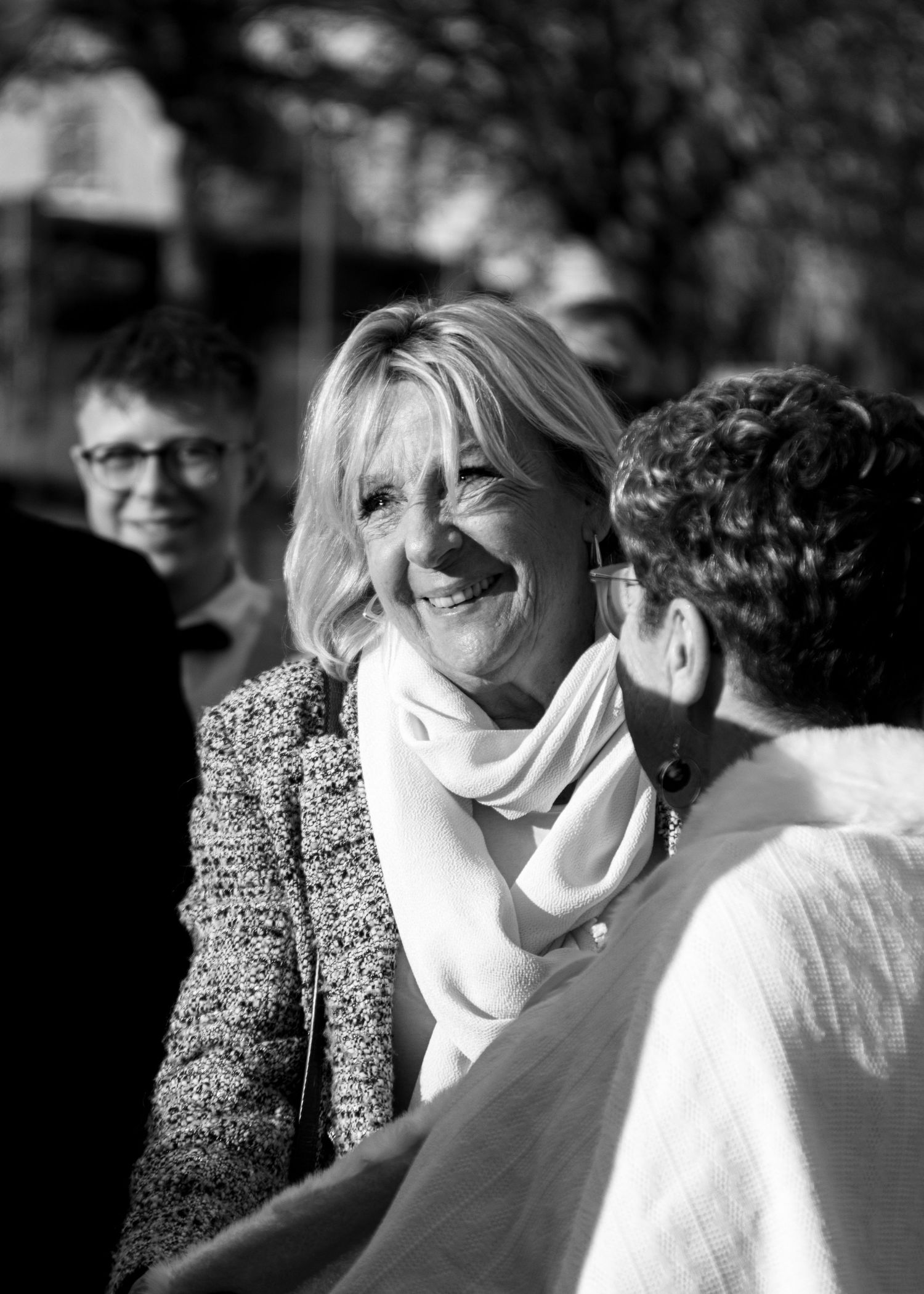 Black and white photo of a smiling blonde woman in a tweed jacket and white scarf greeting people outdoors.