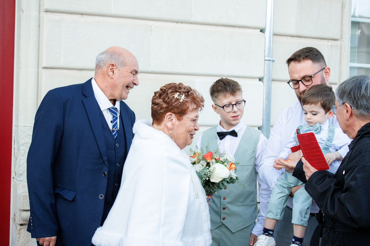 Elderly couple in wedding attire with bouquet, joined by family members including children in formal wear outdoors.