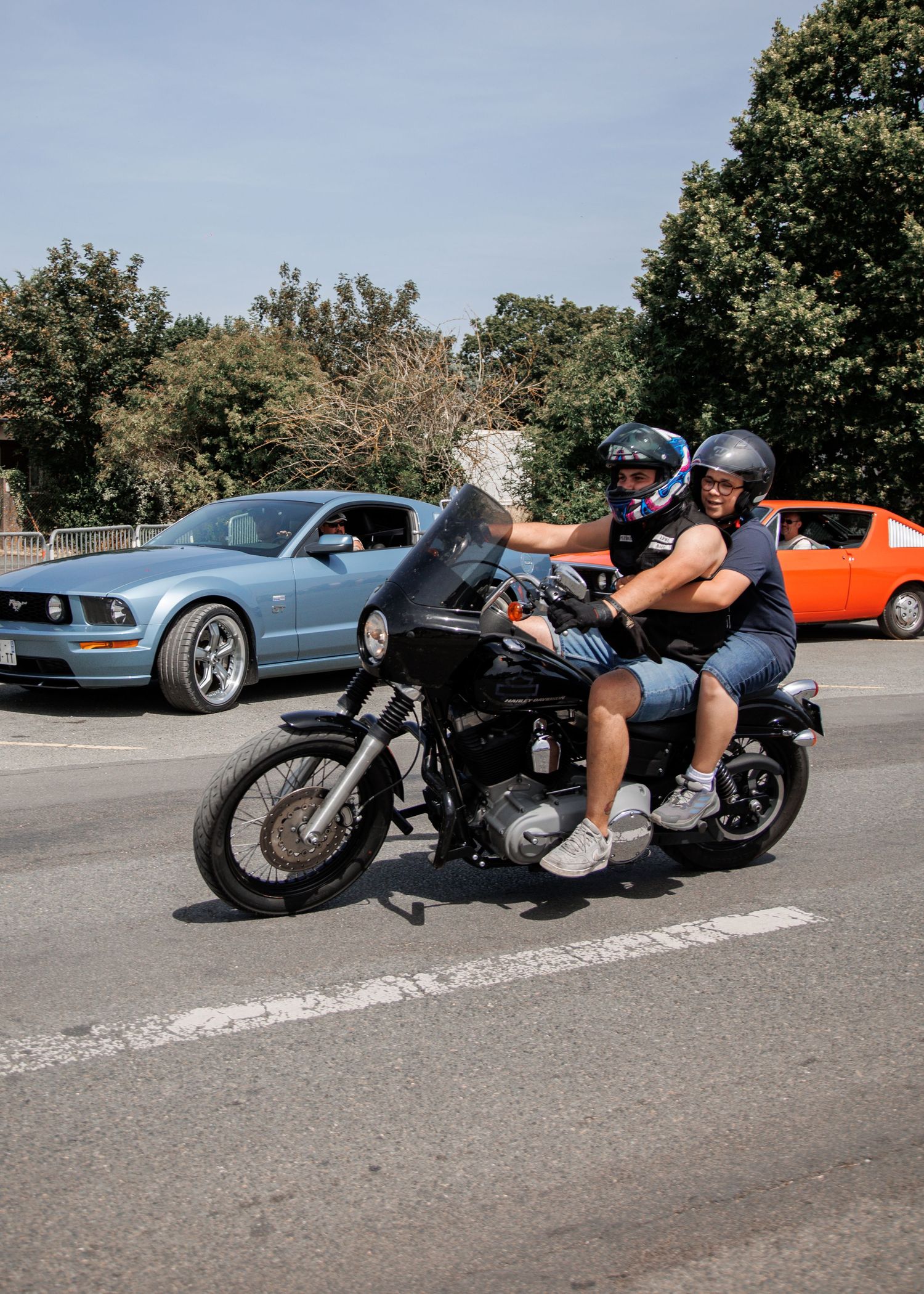 Two riders on a black motorcycle cruise past a blue Ford Mustang and orange car on a sunny tree-lined road.