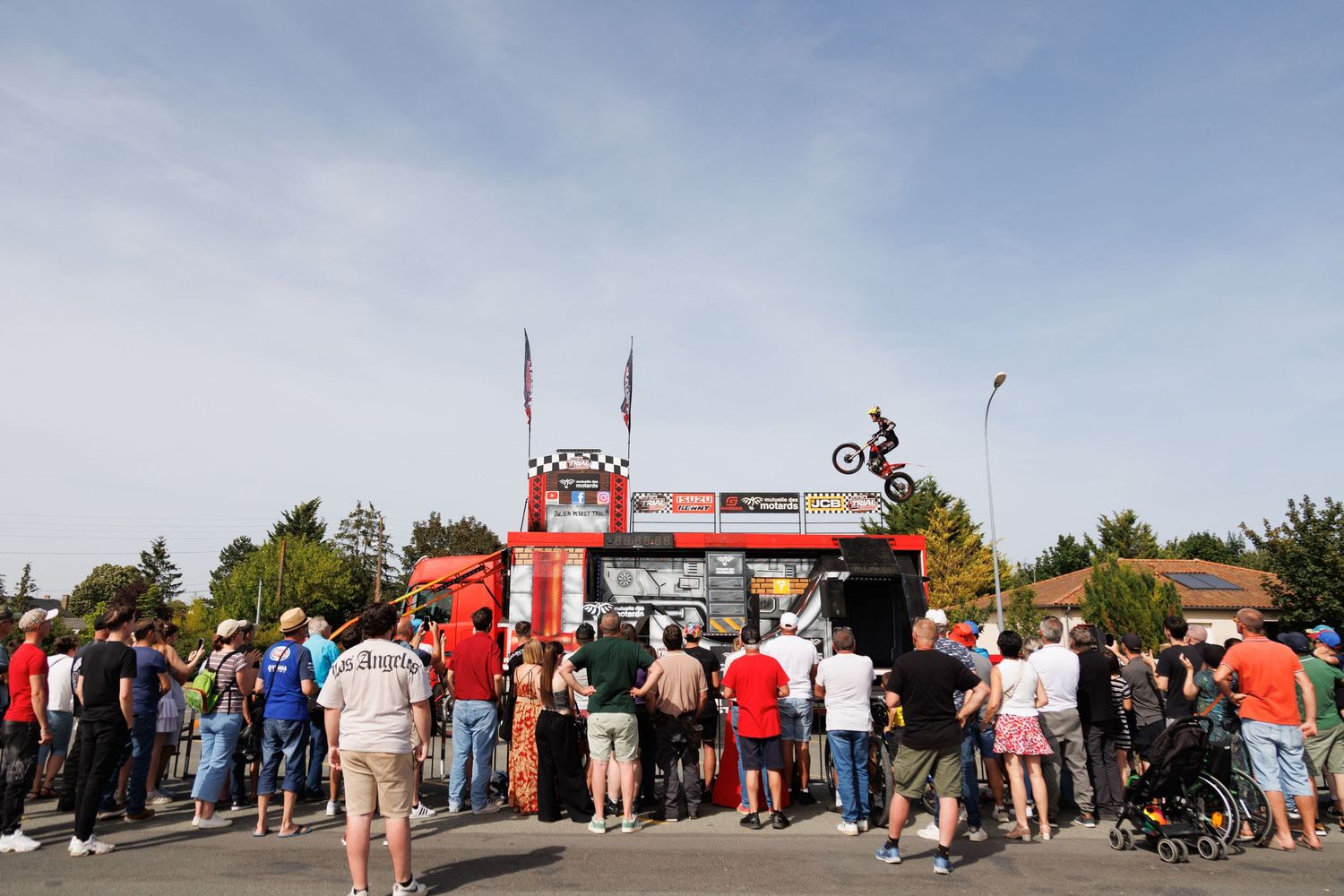 Crowd watches a motorbike stunt rider launch off a ramp at an outdoor motorsport event on a sunny day.