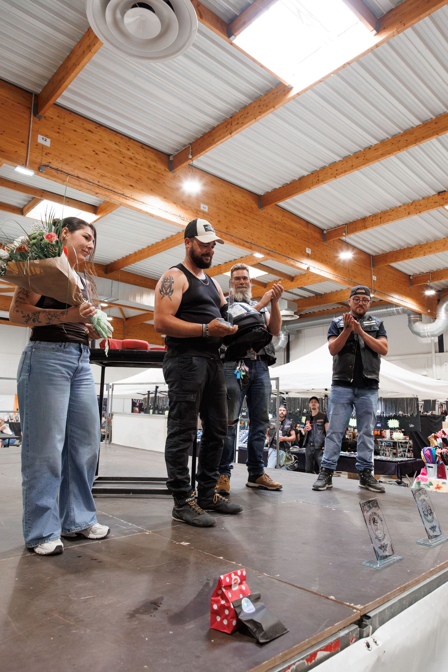 Group of people on stage at an indoor biker event, with a woman holding flowers and men in leather vests clapping.
