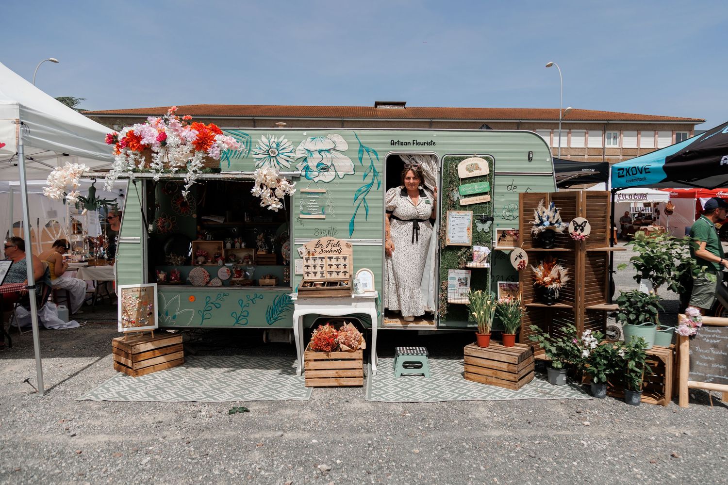 A woman stands in a charming green market stall decorated with flowers and wooden crates at an outdoor bazaar.