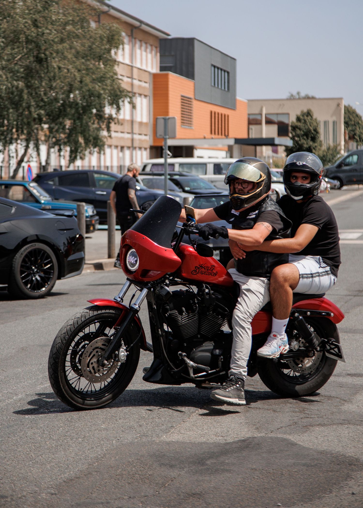 Two riders in helmets on a red motorcycle in a sunny urban parking lot setting.