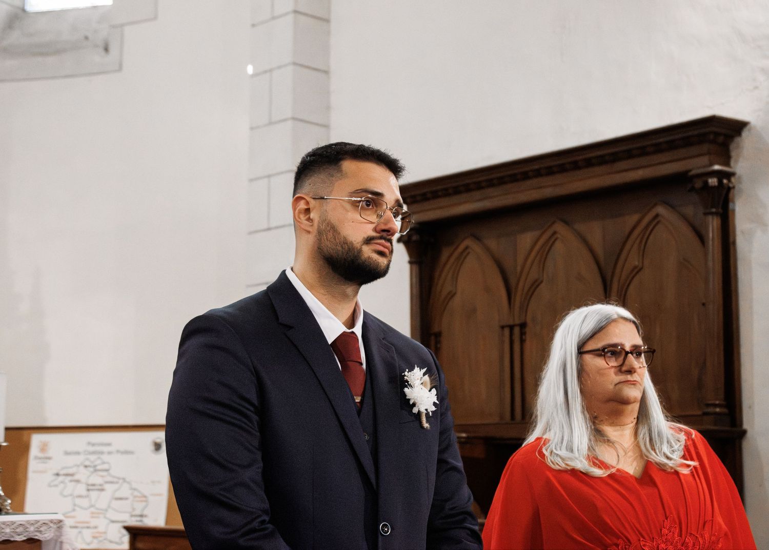 Groom in navy suit with boutonniere stands beside woman in red dress inside a church.