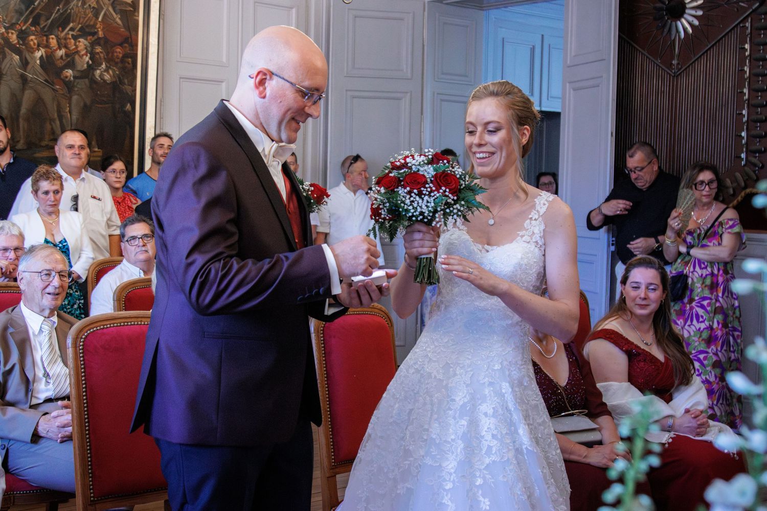 Bride and groom exchanging rings during indoor wedding ceremony, bride holding red bouquet, guests seated in background.