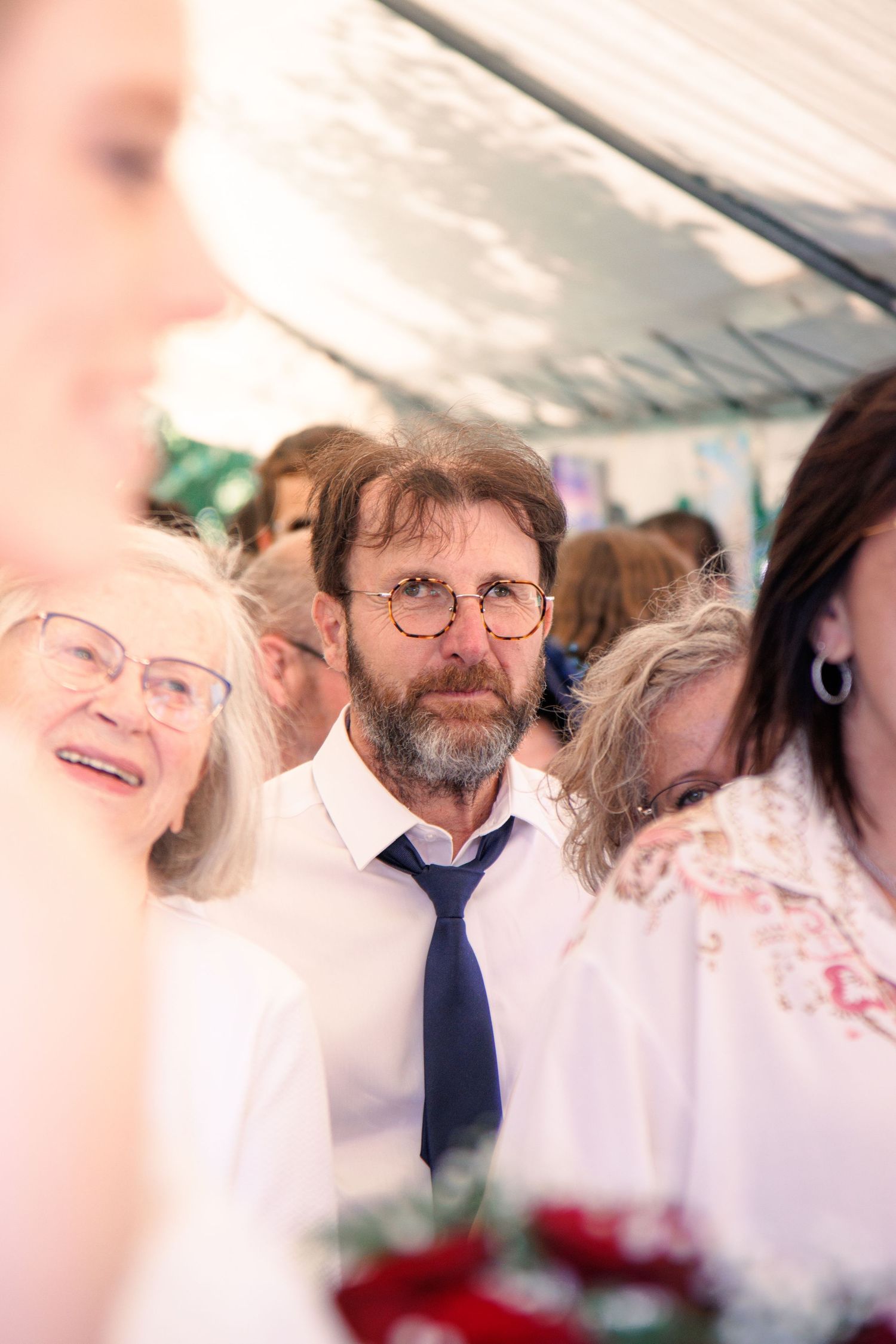 Bearded man wearing glasses and blue tie stands among wedding guests under a decorated tent.