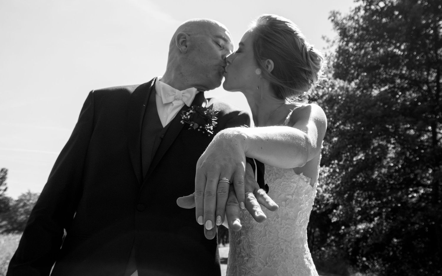 Black and white photo of a bride and groom sharing a kiss, showcasing wedding rings, with trees in the background.