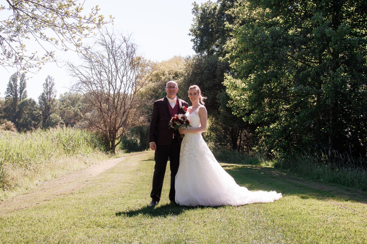 Bride and groom posing together outdoors on green lawn with trees, bride in white gown holding bouquet.