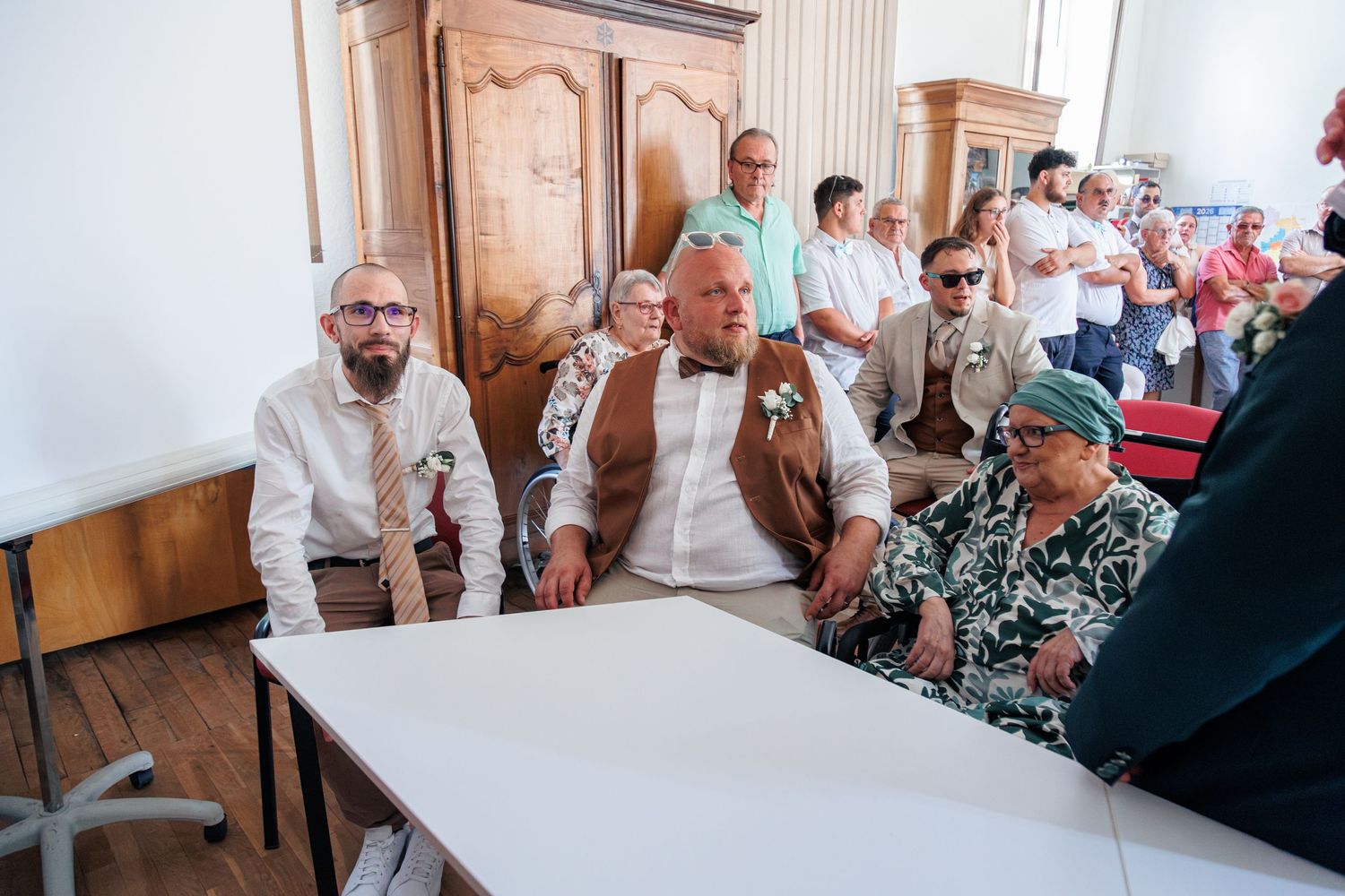 Wedding guests seated indoors at a formal ceremony, with a large man in a green bow tie as the focal point.