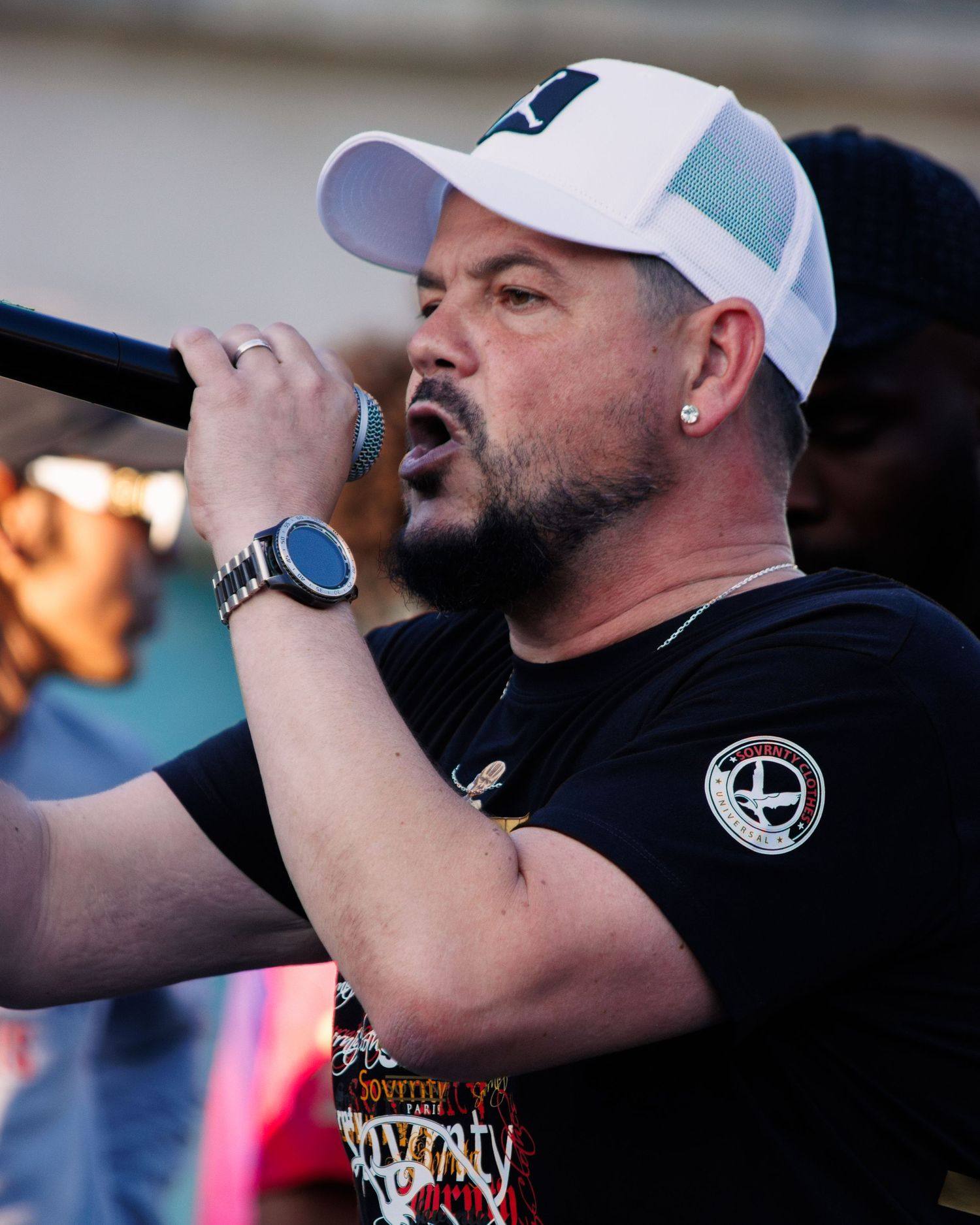 Performer in white baseball cap and black t-shirt singing passionately into microphone during outdoor performance.