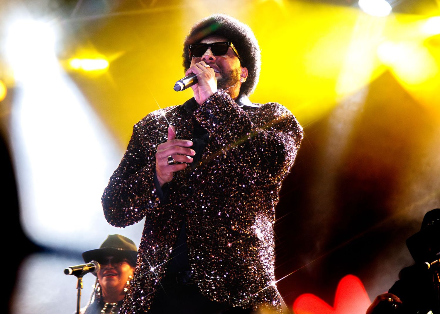 Performer in sparkly jacket sings on stage with bright yellow concert lights glowing behind them.