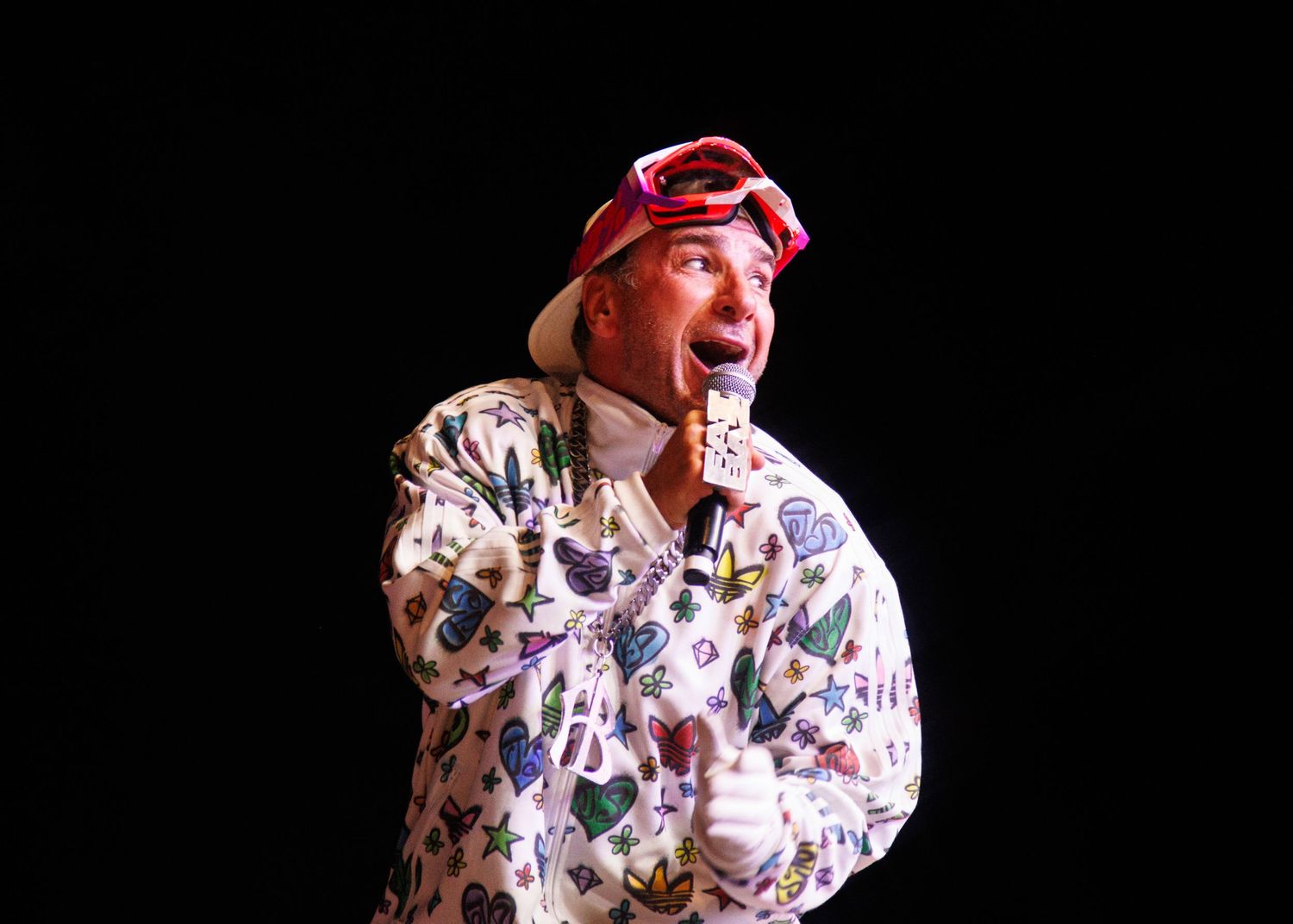 Person wearing a red headpiece and patterned jacket performs expressively on a dark stage.