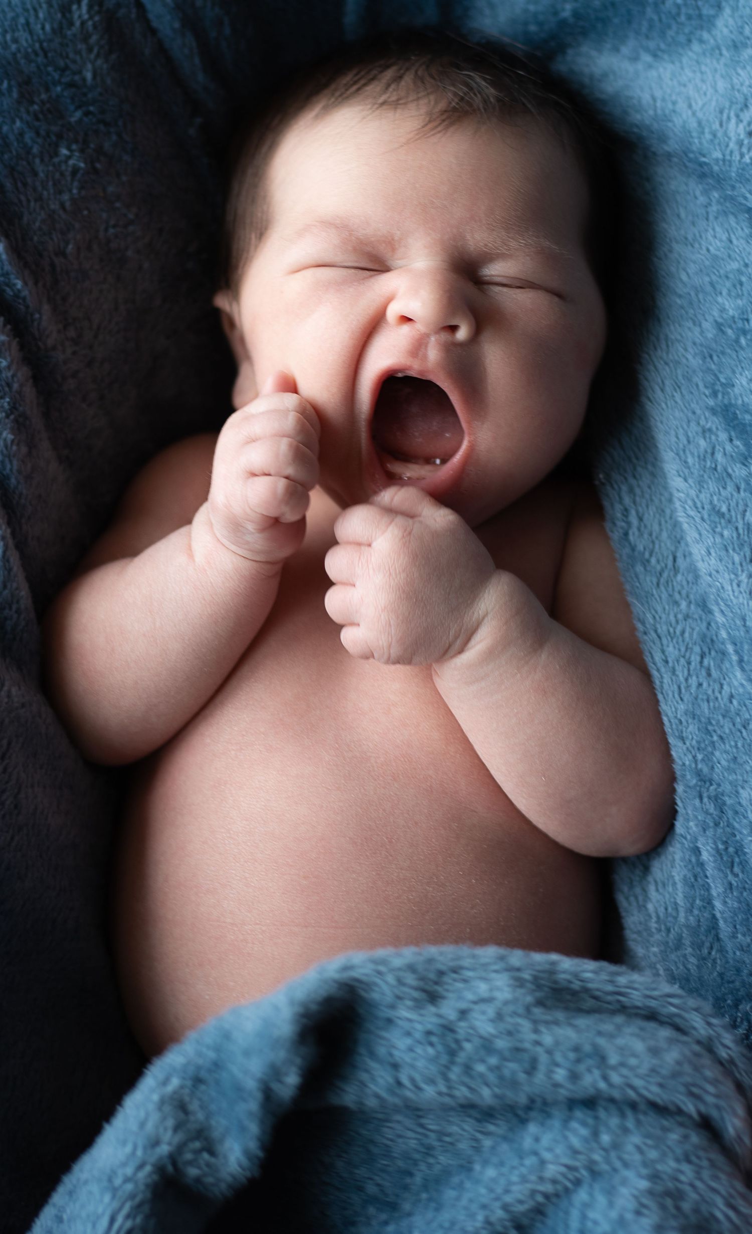 A yawning baby lies on blue blankets during a newborn photo session.