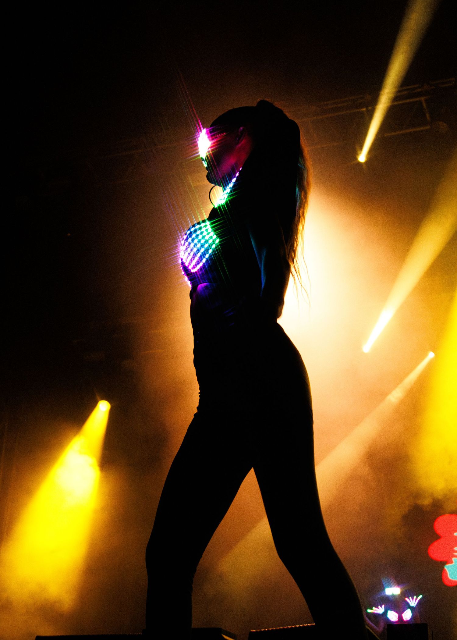 Silhouetted dancer in colorful neon lights performs on dark stage with yellow spotlights in background.