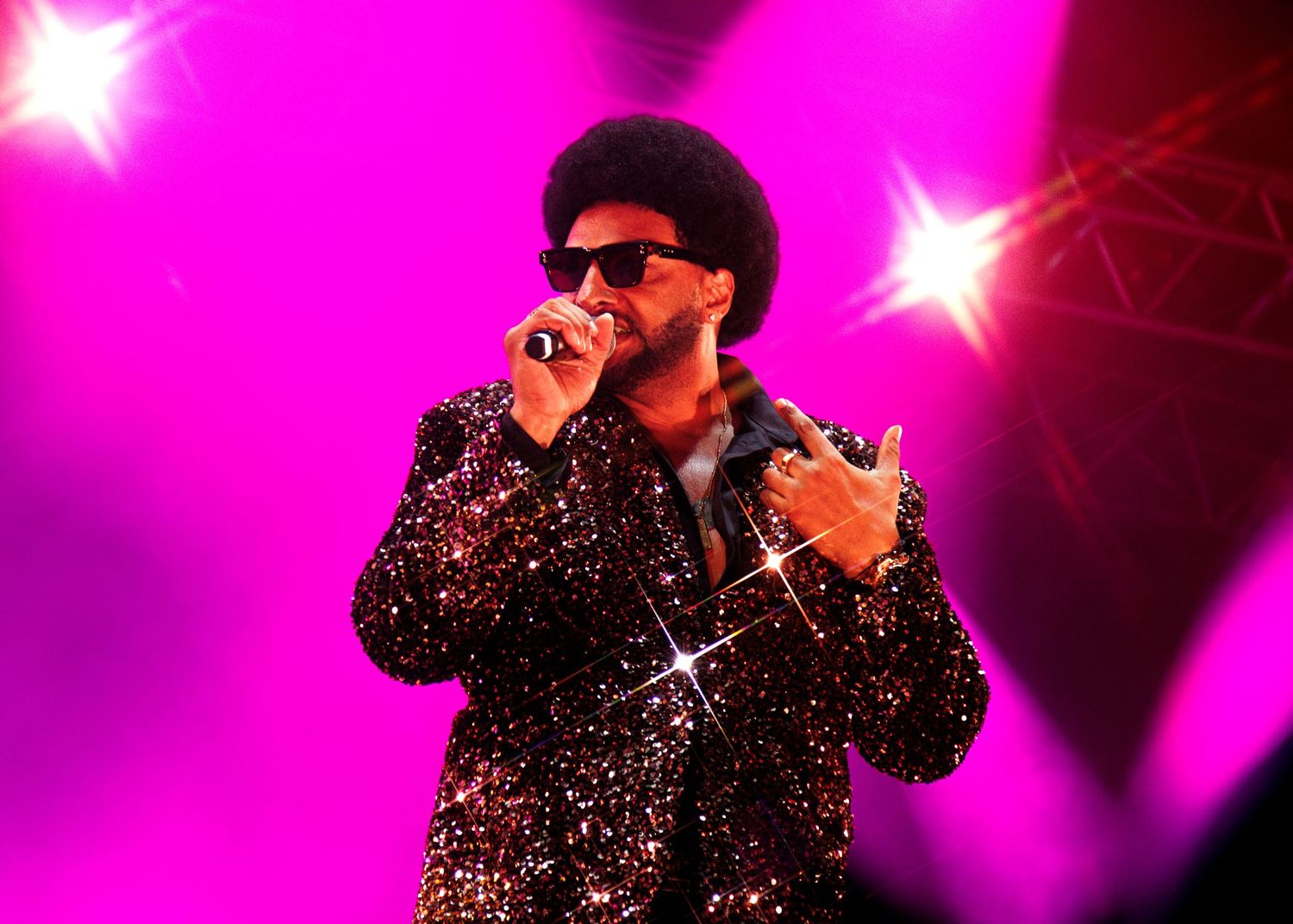 A performer in a sparkly black outfit and sunglasses poses dramatically on stage with bright pink lighting effects.