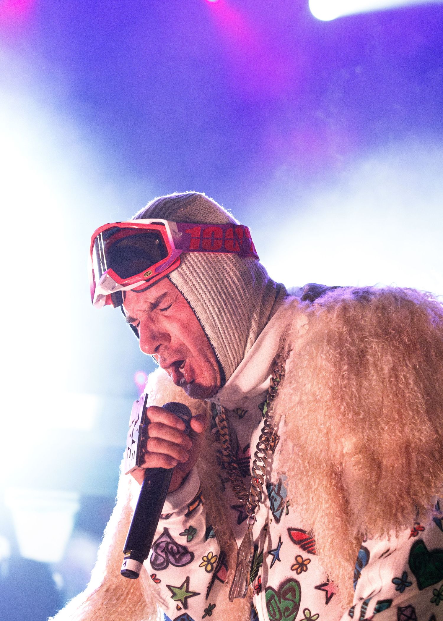 Performer in red ski goggles and white knit cap singing into microphone under purple stage lighting.