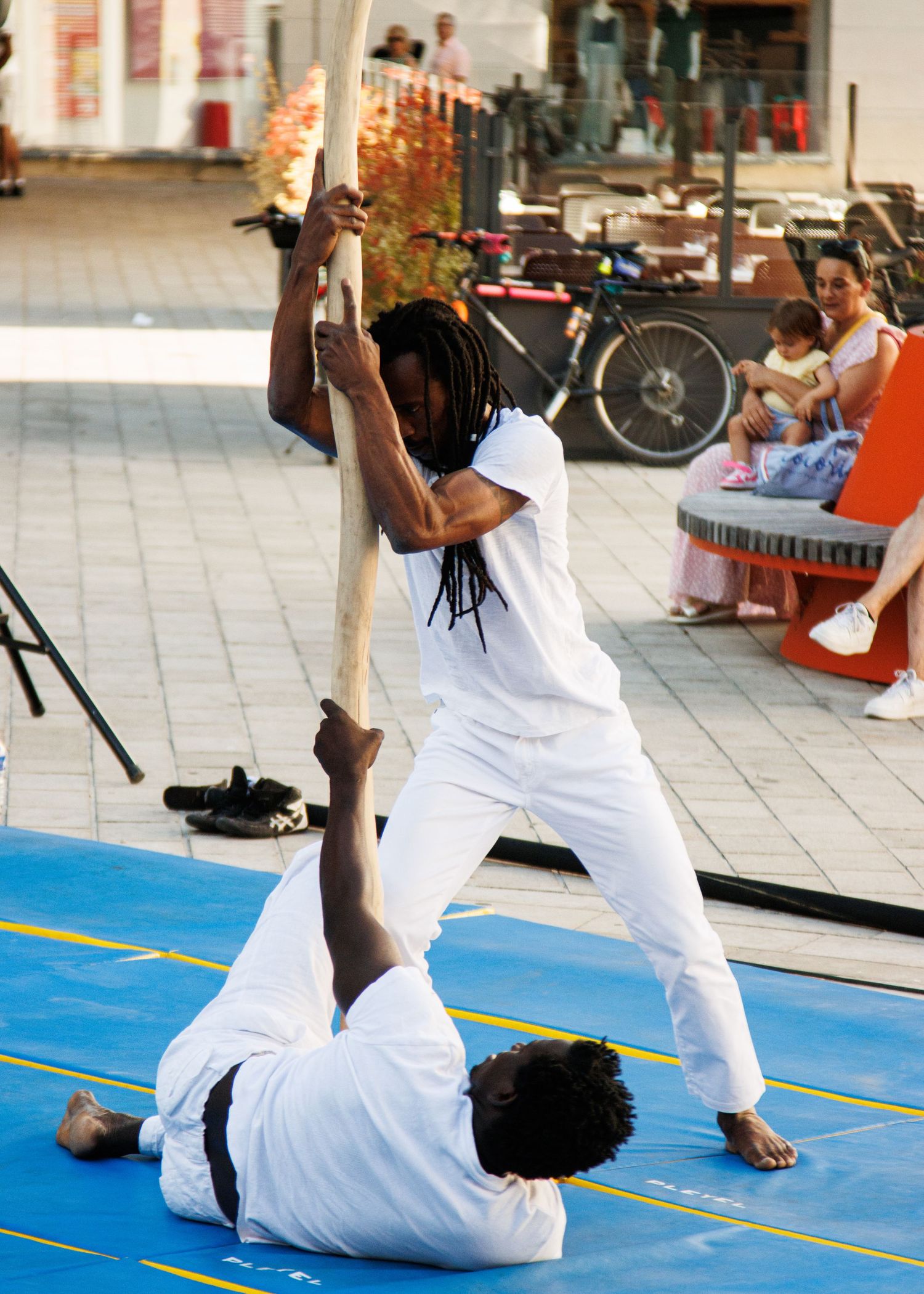 Street performers demonstrate an advanced acrobatic move on a blue mat in an urban setting.