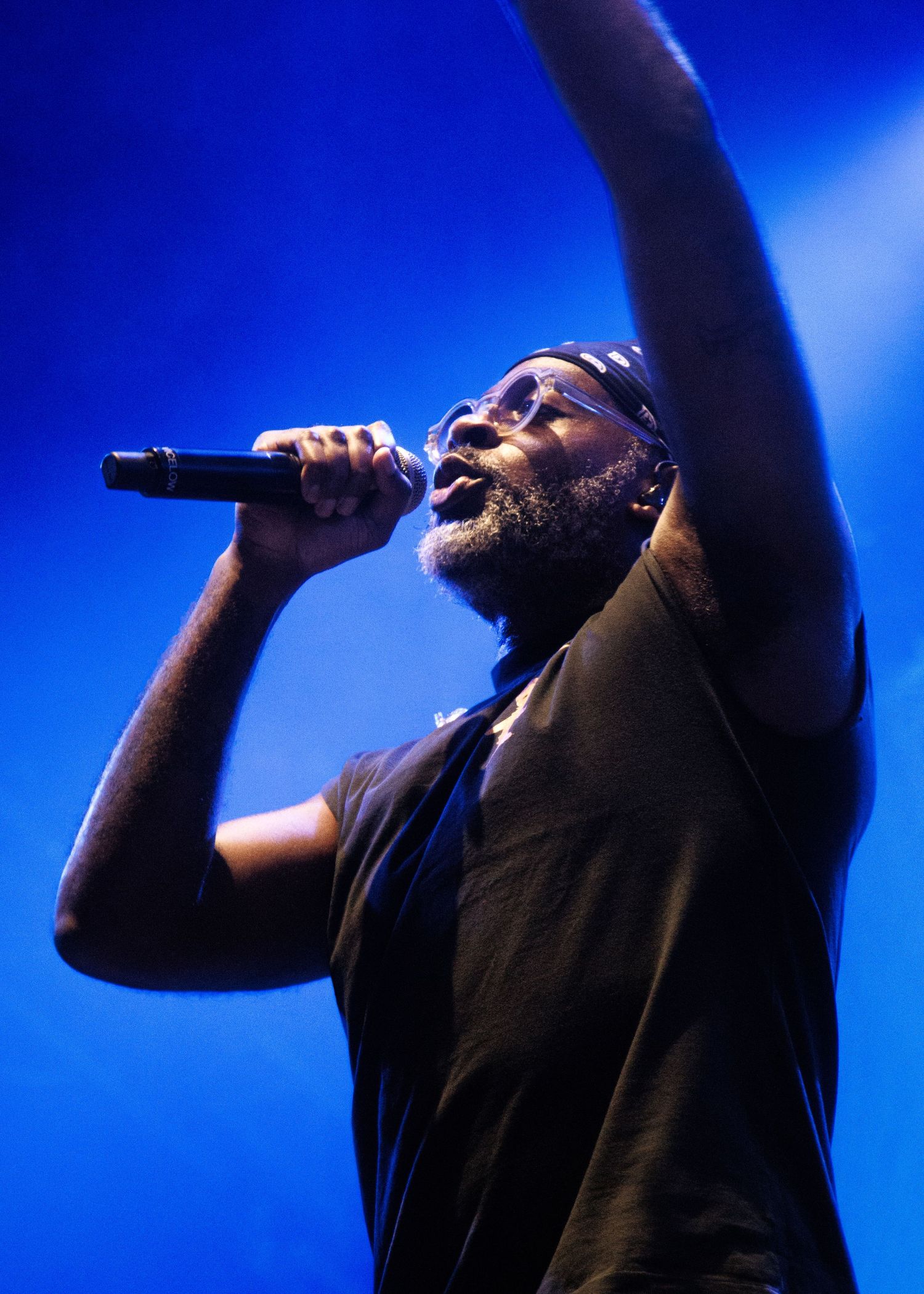 A performer in a black t-shirt sings passionately on stage against a vibrant blue backdrop during a concert performance.