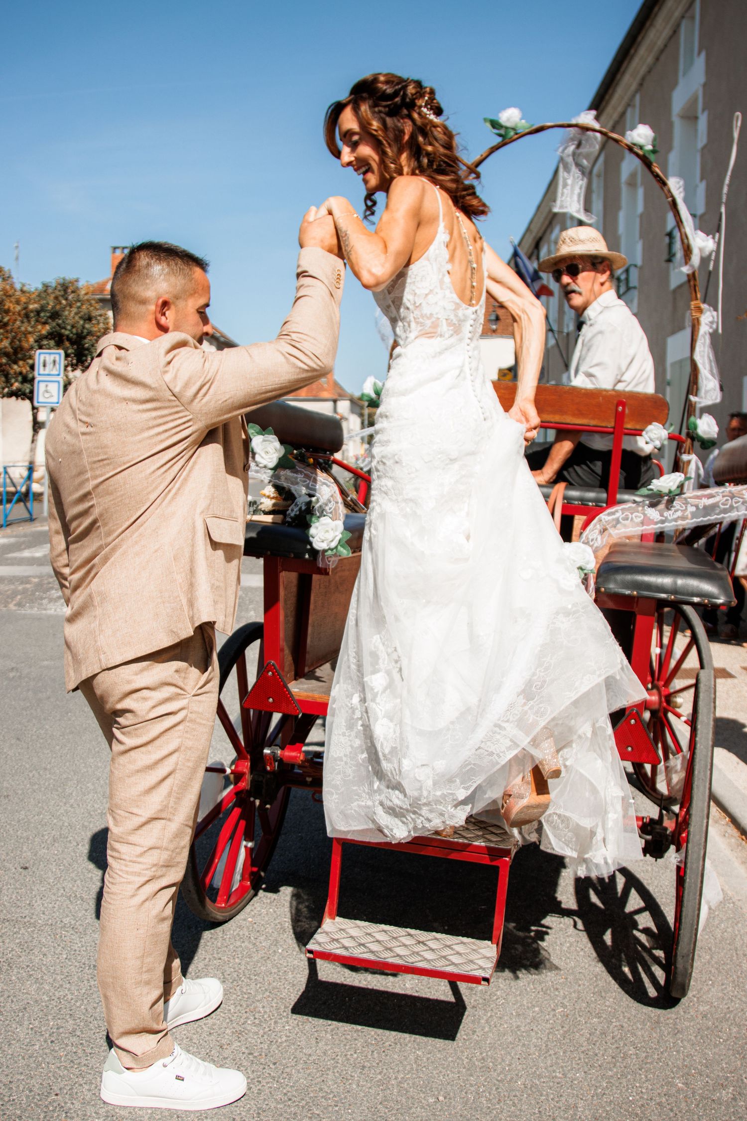 A couple in wedding attire poses playfully with a red vintage wagon on their special day.