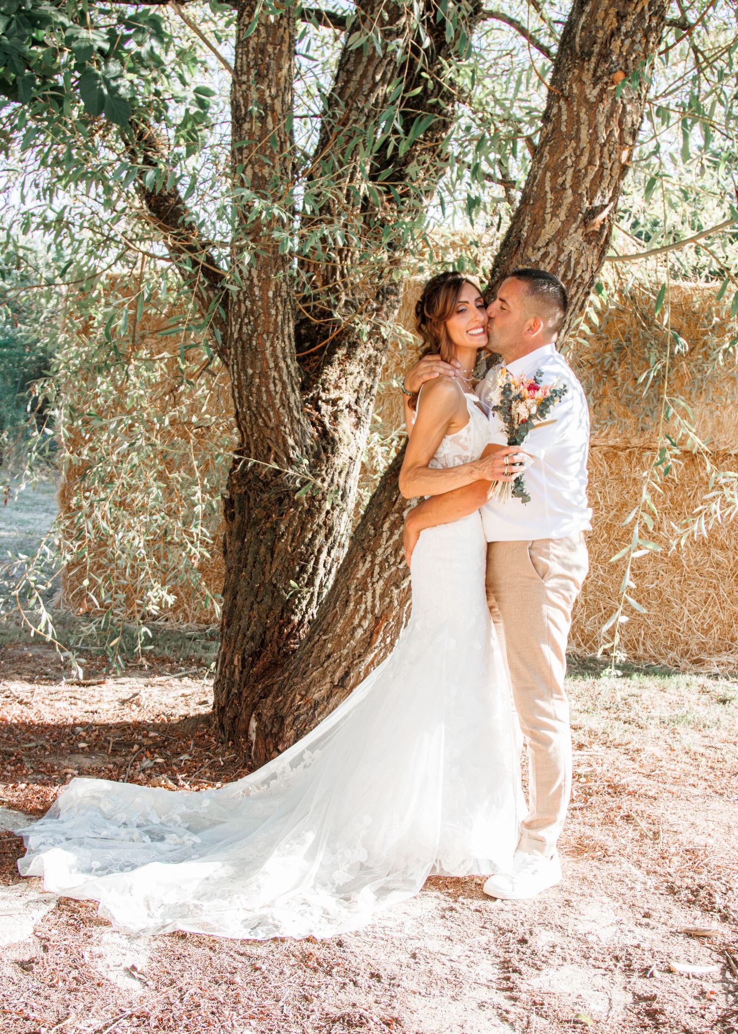 A romantic wedding couple shares intimate kisses under a large tree in an outdoor setting.