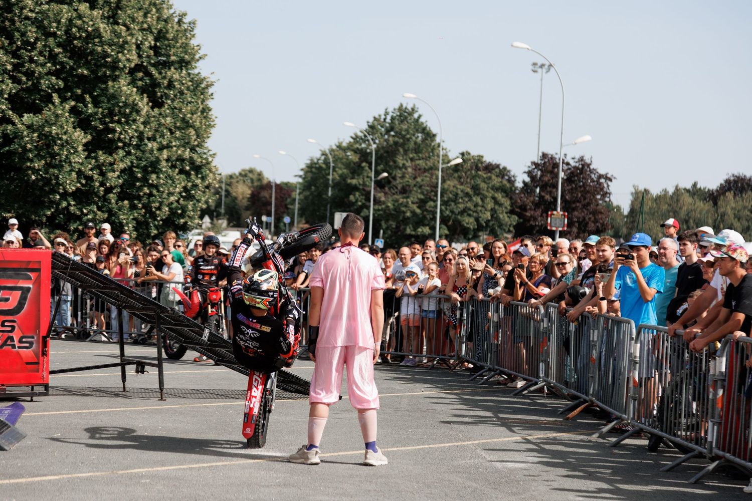 Stunt performer in pink outfit with motorcycle on ramp entertains large crowd at outdoor event on sunny day.