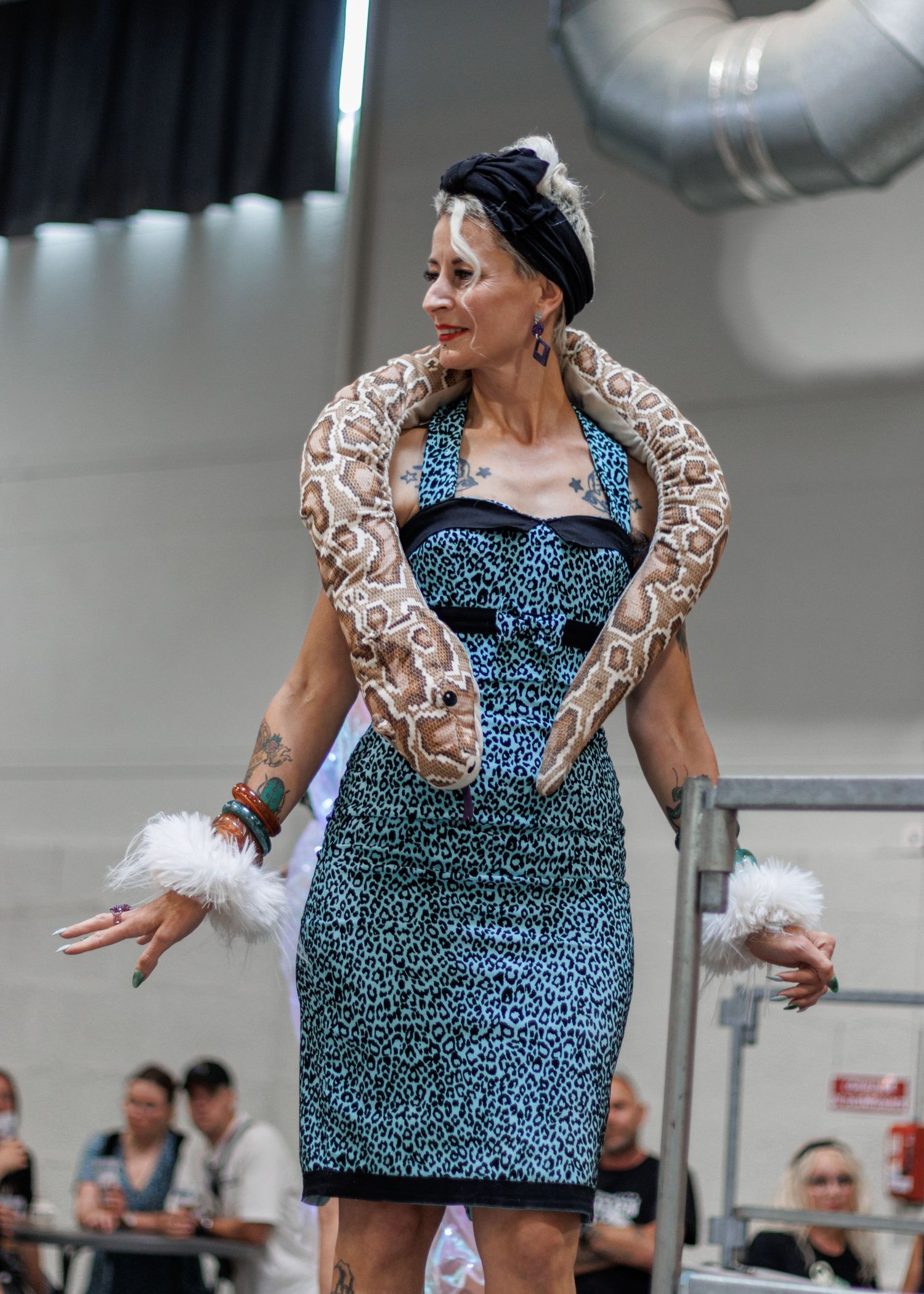 Woman in blue leopard dress with large boa constrictor draped over shoulders, wearing white feather wrist accessories.