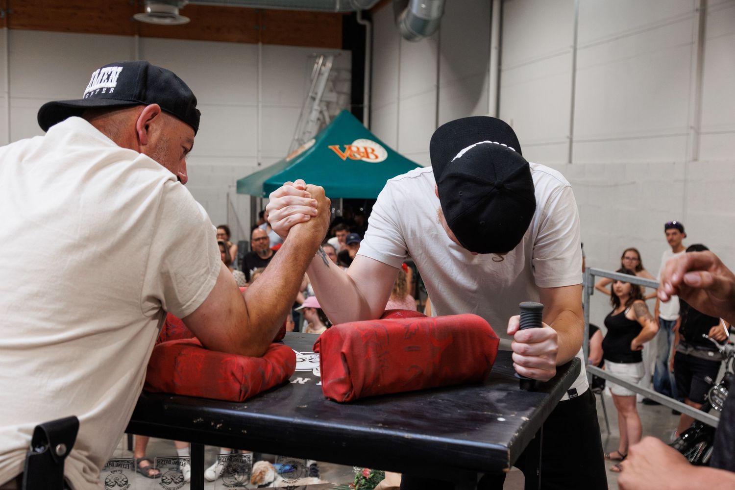 Two men competing in an arm wrestling match at an indoor event with a crowd watching in the background.