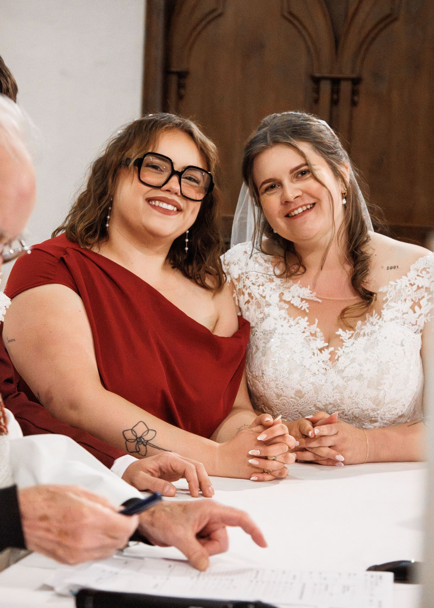 Two brides smile while signing wedding documents, one in lace gown with veil, other in red dress with novelty glasses.