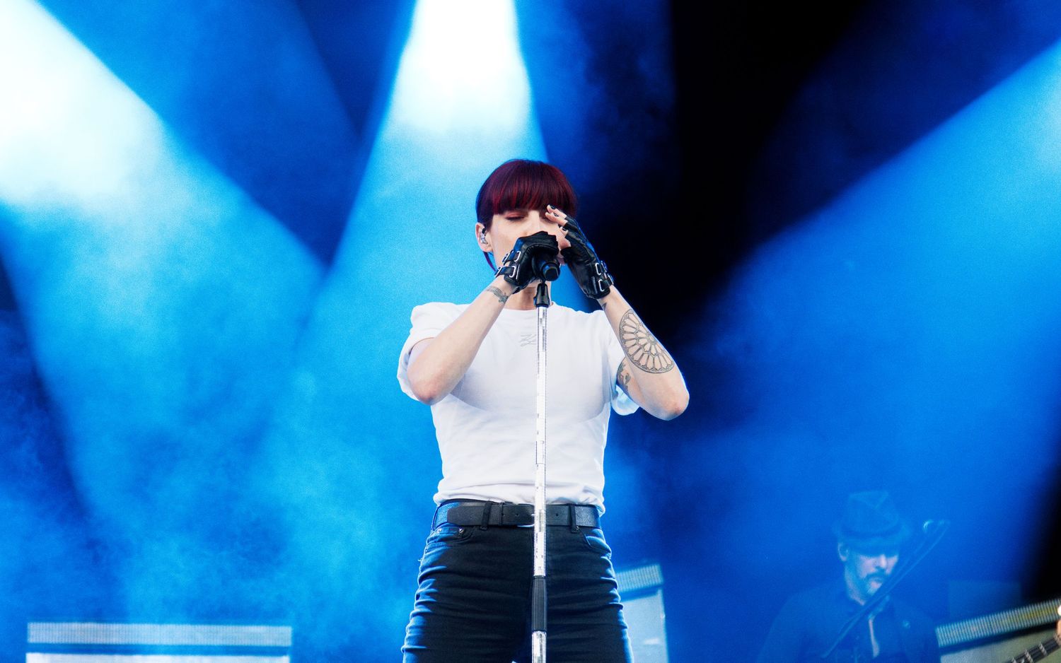 Female singer performing on stage with blue stage lights, wearing white top and black pants, holding microphone.