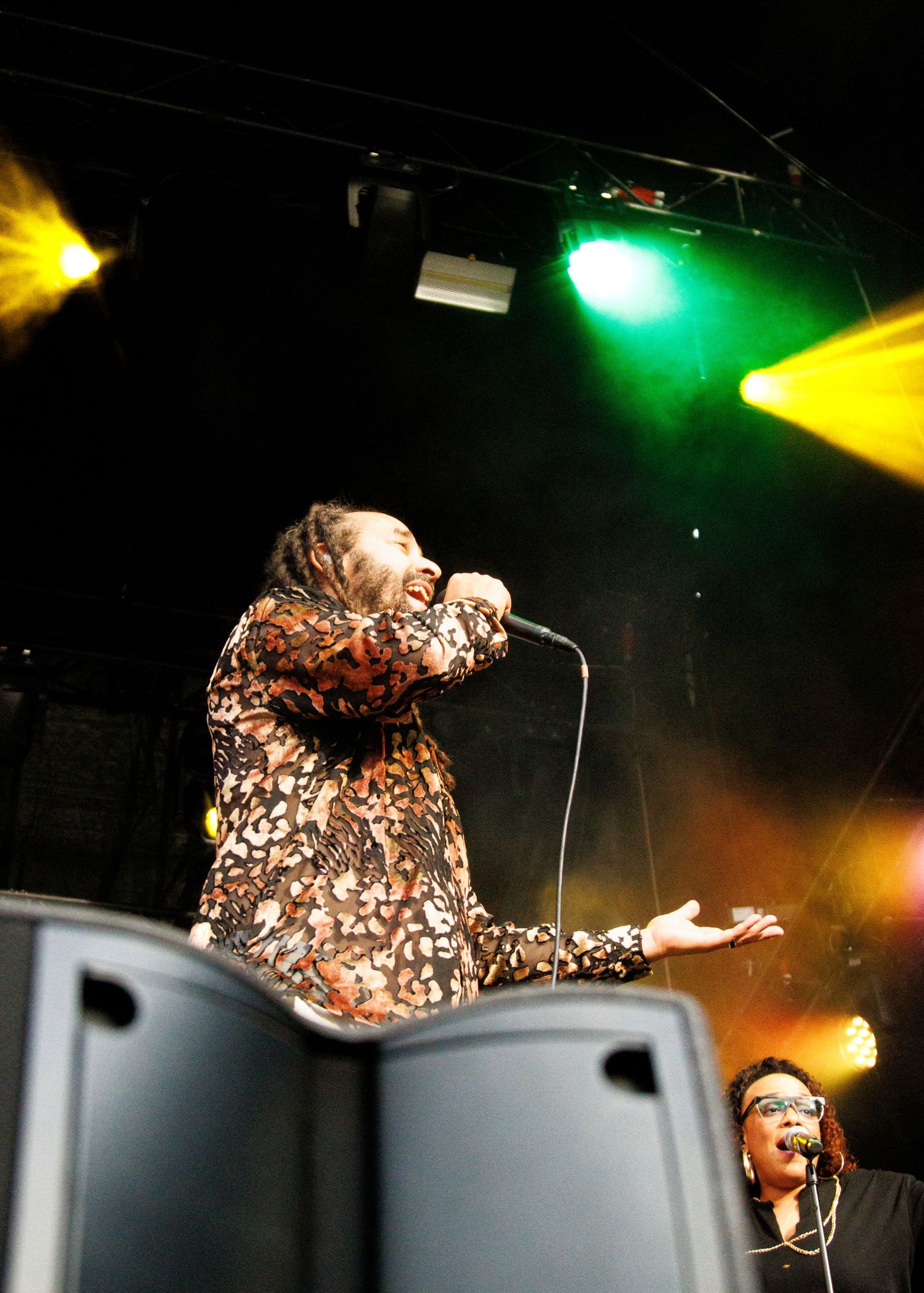 Singer performing on stage under colorful green and yellow lights, wearing a floral outfit and holding a microphone.