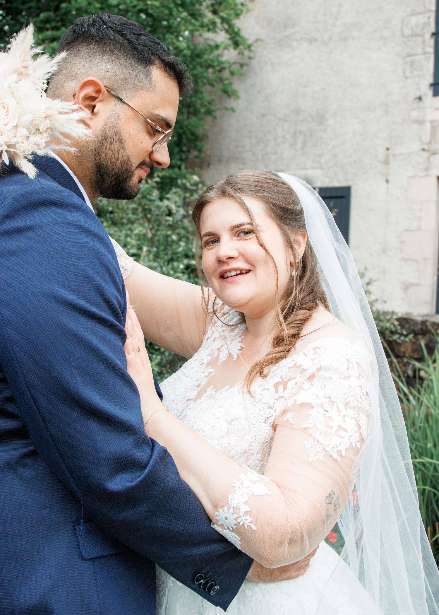 A bride in lace gown and veil smiles at groom in navy suit during outdoor wedding portrait.