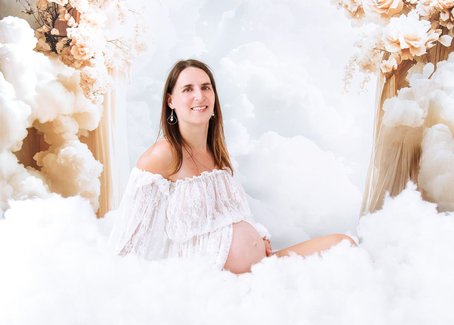 Ethereal maternity photo with soft white lace dress surrounded by dreamy clouds and warm autumn-toned floral decorations.