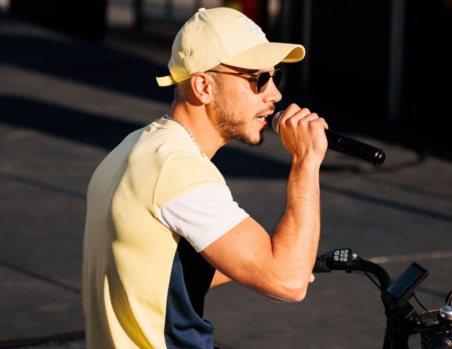 Performer in yellow shirt and baseball cap singing into microphone during outdoor concert.