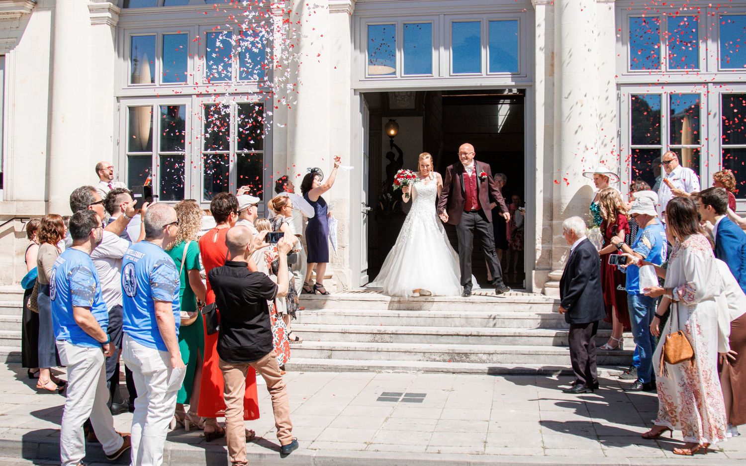 Newlyweds exit building as guests throw confetti during outdoor wedding celebration.