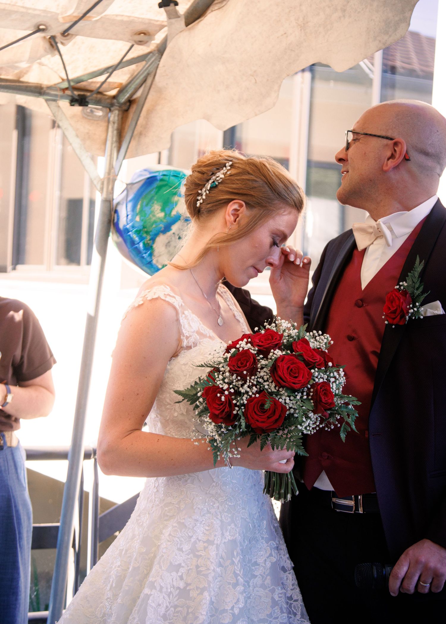 Emotional bride in lace gown wiping tears holding red rose bouquet beside smiling groom.