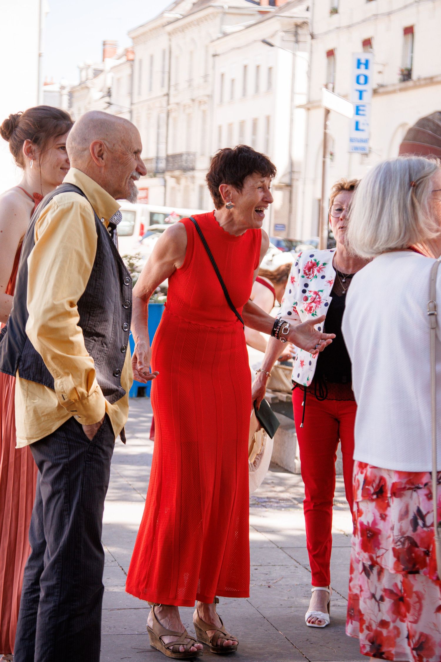 A woman in a red dress socializes with a group of people on a sunny street in France.