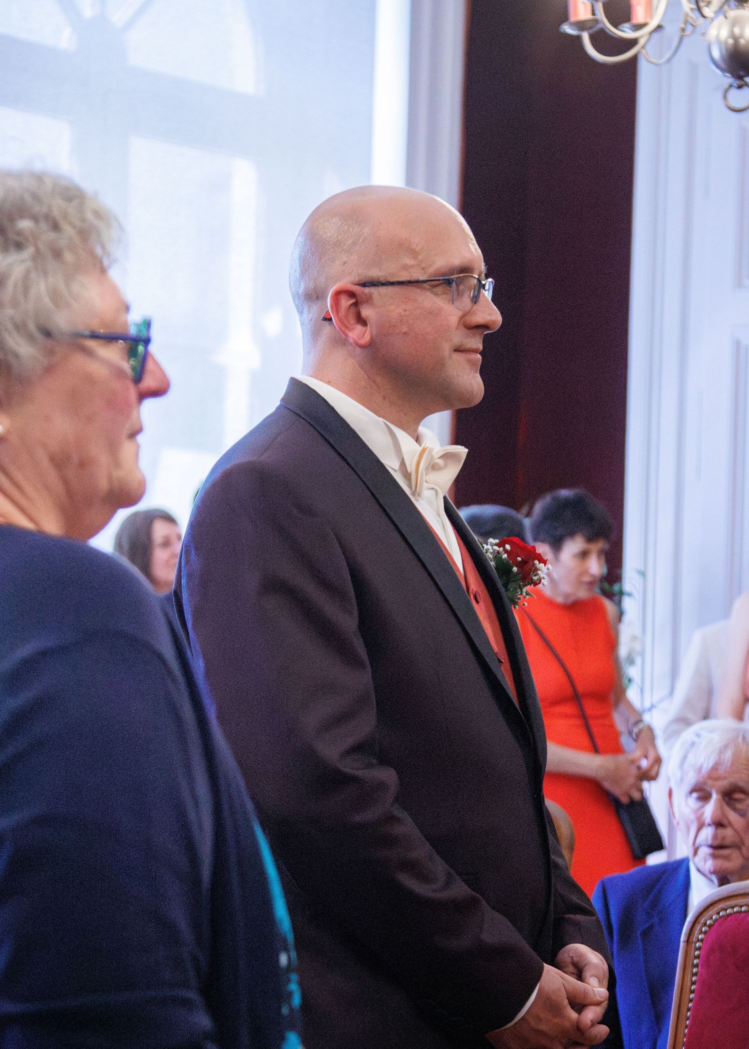 A bald man in a dark suit with boutonniere stands at a wedding ceremony indoors.