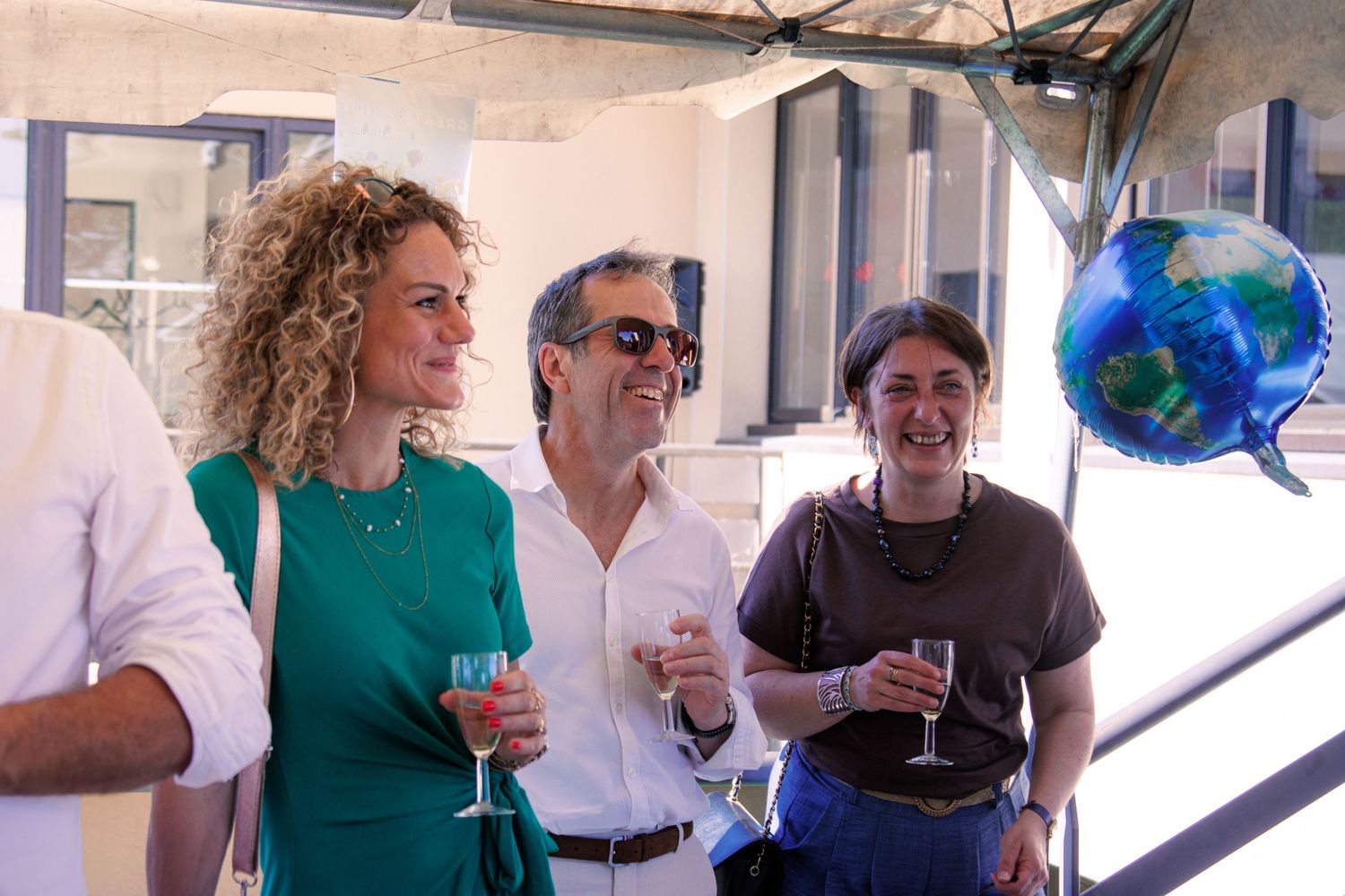 Three guests smiling and holding champagne glasses at outdoor celebration with decorative globe balloon in background.