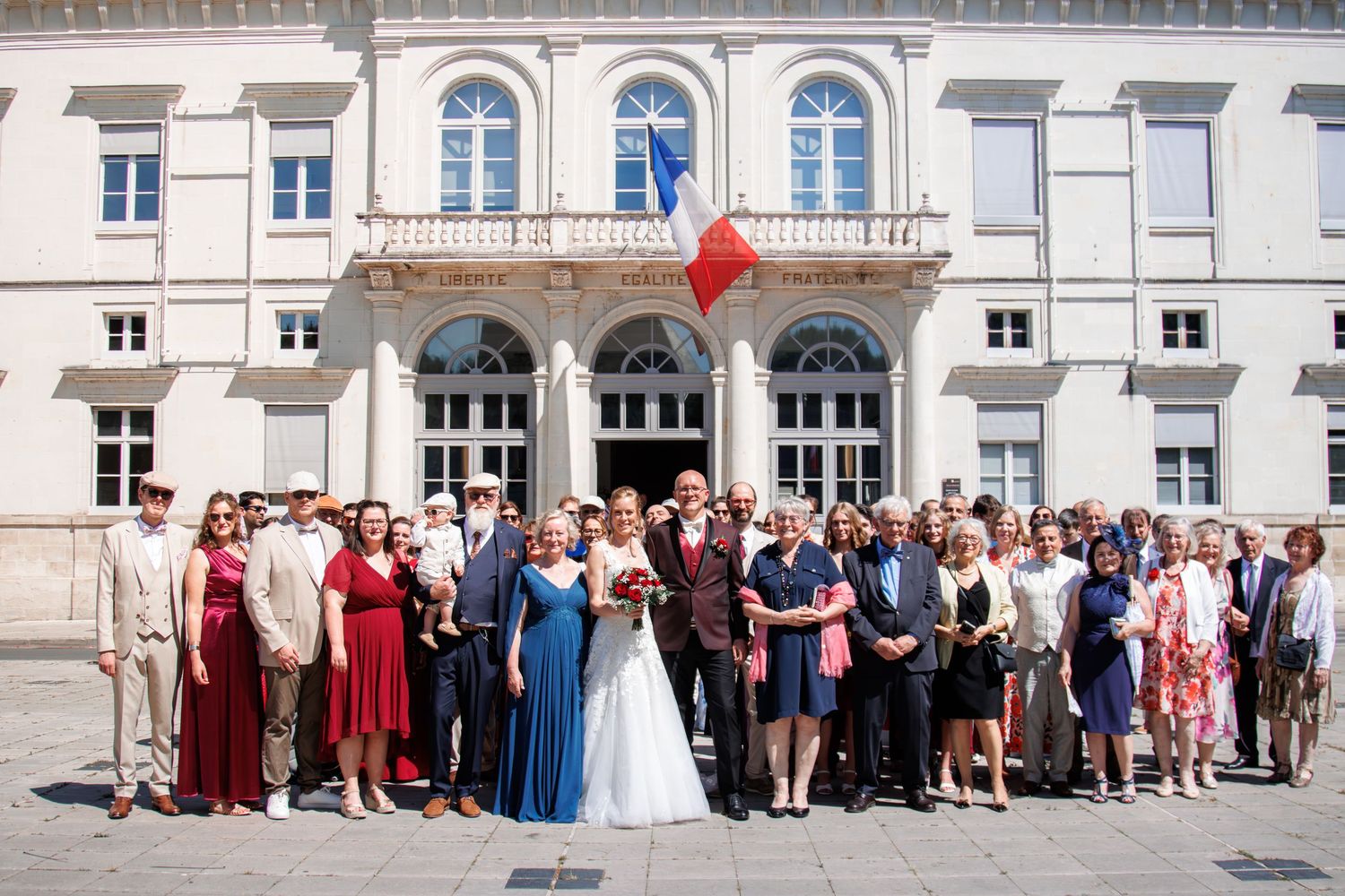 Large wedding party group photo in front of a classical French building with a French flag displayed.