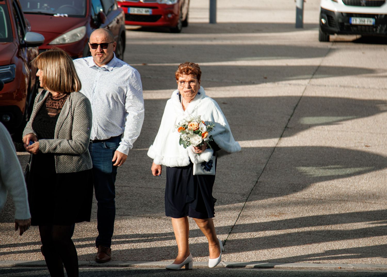 Elderly woman holding bouquet walks with man in white shirt across parking lot in casual outdoor setting.