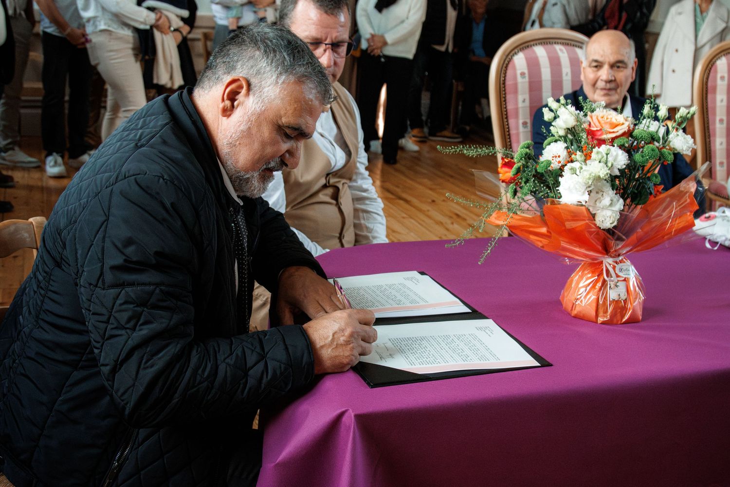 Man in black jacket signing official documents at purple table with flower bouquet and guest in background.