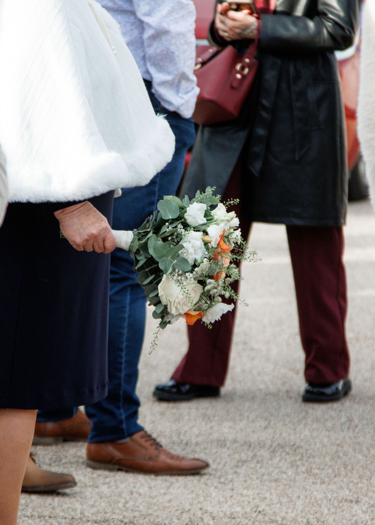 A bride holding a white and green wedding bouquet with orange accents while guests stand nearby outdoors.